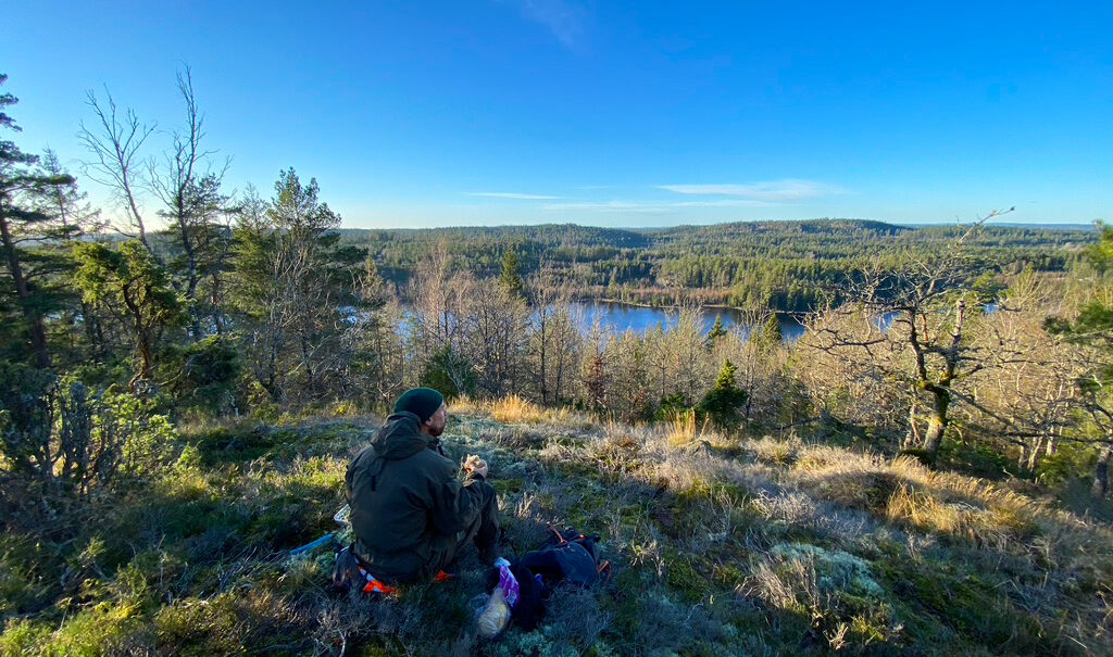 Person sitter och fikar ute i skogen och ser på sjöutsikten. Foto.